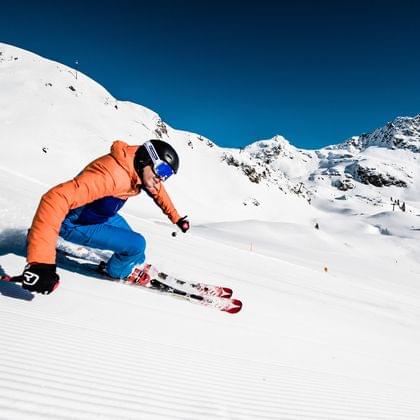 Skier in orange jacket carving on groomed slope in alpine ski resort with mountain backdrop